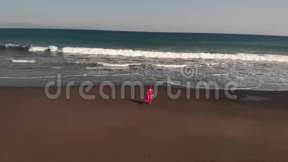4巴厘岛黑色火山海滩上年轻女子穿着粉红色连衣裙行走的空中飞行镜头视频的预览图