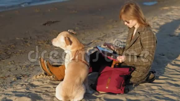 河边海滩上的一位年轻女子用电脑平板电脑喂了一只棕色的龙虾狗春天或寒冷的夏天视频的预览图