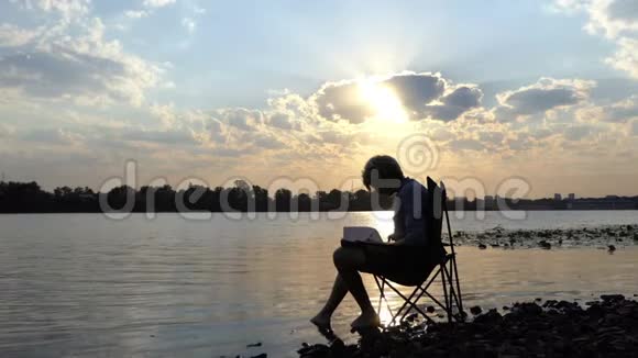 作家在斯隆莫夕阳下的河岸上的啜泣阅读和写下他的笔记视频的预览图