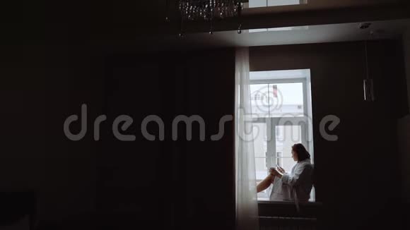 一个穿着白色外套的悲伤女人端着一杯茶坐在窗台上视频的预览图