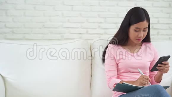 快乐的年轻亚洲女人在家学习在手机上看短信或短信微笑着坐在沙发上视频的预览图
