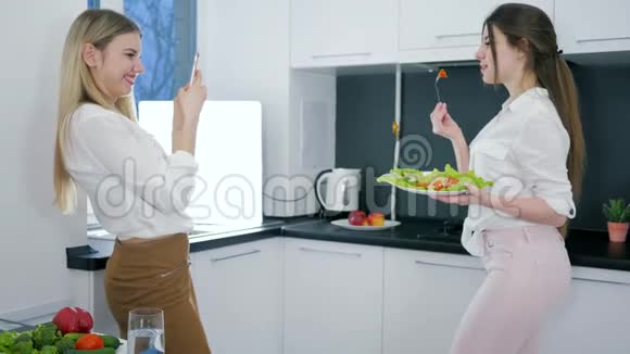 漂亮的女朋友在厨房里使用智能手机女孩用叉子和沙拉摆姿势视频的预览图