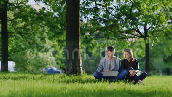 年轻可爱的一对享受笔记本电脑坐在公园的草地上树下视频的预览图