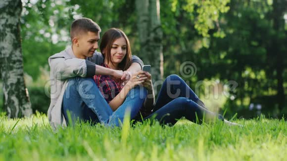 男人和女人拥抱坐在公园的草地上看着手机屏幕视频的预览图