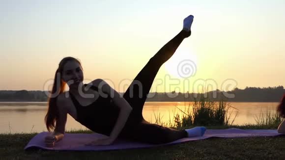 孕妇躺在垫子上抬起她的腿在日落的湖岸上视频的预览图