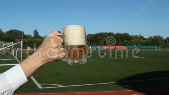 一个男人手拿着一杯啤酒背景是一个足球场在那里他们踢足球特写镜头视频的预览图