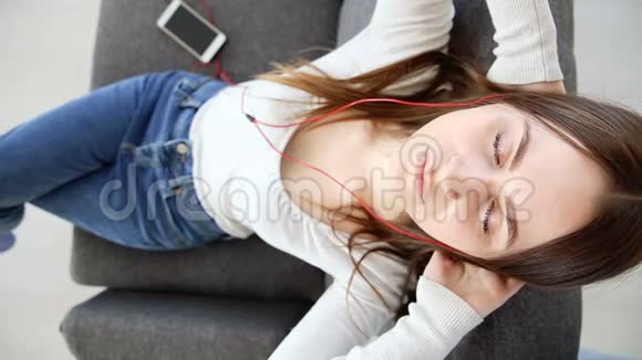 漂亮的女孩在家里沙发上用耳机听音乐视频的预览图