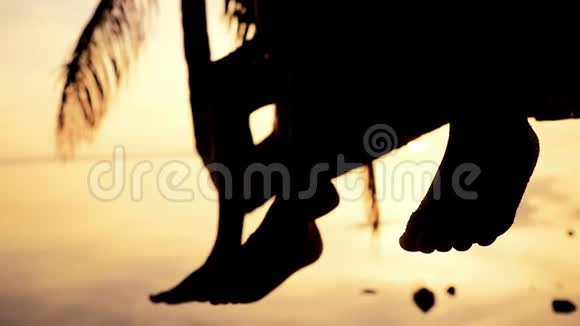 在令人惊叹的日落中年轻快乐的父亲和他的女儿坐在棕榈树上的剪影挥舞着双腿关门视频的预览图