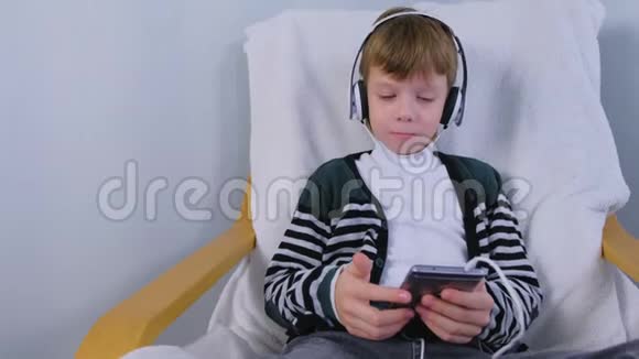 男孩正坐在家里的扶手椅上用智能手机耳机听音乐视频的预览图