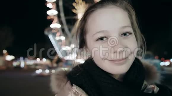 一个快乐的年轻女孩的特写肖像她在夜晚发光的环形交叉路口的背景下看着摄像机视频的预览图
