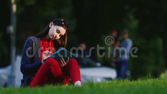 时髦的女人坐在公园的草地上享受着平板电脑在背景下人们走路视频的预览图