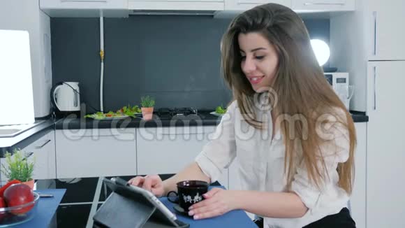 早晨黑发女人在餐桌上用平板电脑在家里的厨房里喝茶视频的预览图