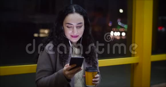 漂亮的女人坐在公共汽车站用智能手机喝咖啡在后台有夜城视频的预览图