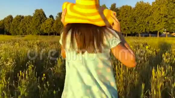 美丽的女孩戴着黄色的帽子在日落的天空上的绿色麦田上奔跑自由概念日落时的麦田慢慢视频的预览图
