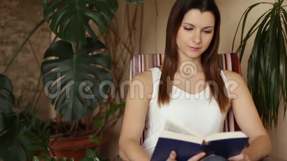 年轻迷人的女人在客厅里坐在舒适的椅子上读有趣的书微笑的时候视频的预览图