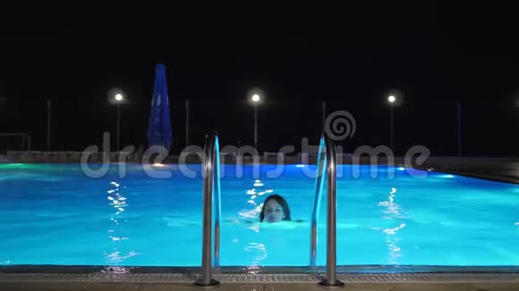 穿着泳衣的年轻女子从彩色的水中出来抱着游泳池的梯子进入傍晚视频的预览图