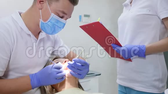 正畸医生用牙具看着女人的嘴旁边的护士在门诊记事本上写诊断视频的预览图