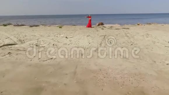 穿着红色长裙翩翩起舞的年轻姑娘举起双手站在蓝色海水和壮观景色附近的大石头上视频的预览图