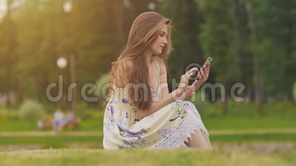 年轻迷人的夏装女子手里拿着手机坐在绿草上一个公园夏天娱乐视频的预览图