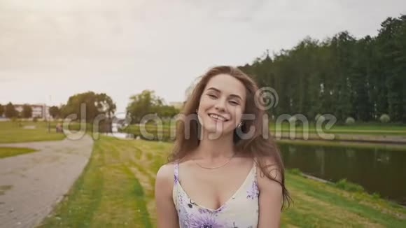 美丽的年轻女孩长卷发穿着一件浅色连衣裙沿着池塘去公园她高兴地摆姿势视频的预览图