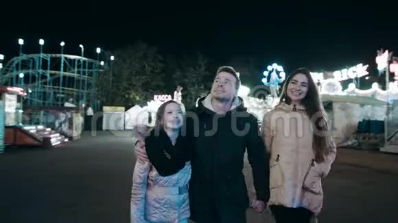 一个美丽的年轻家庭晚上在游乐园里散步在明亮的景点中视频的预览图