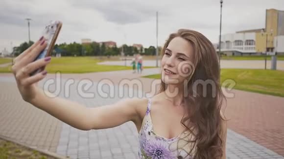美丽迷人的年轻女孩长发和着装正在做照片拍摄使用智能手机在城市公园视频的预览图