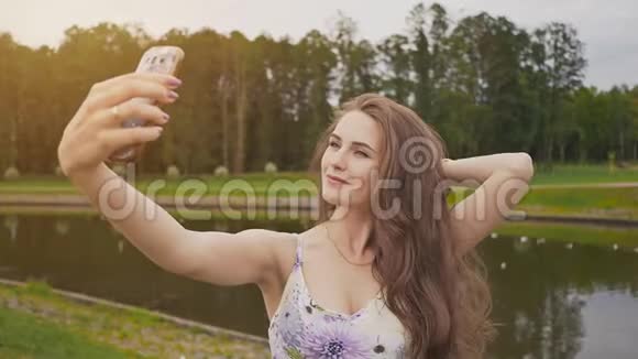 美丽迷人的年轻女孩长发和着装正在做照片拍摄使用智能手机在城市公园视频的预览图