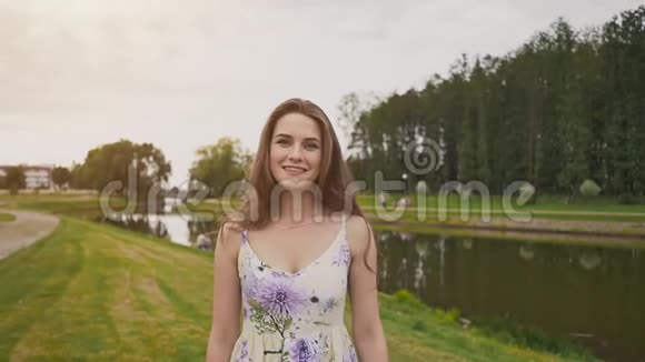 美丽的年轻女孩长卷发穿着一件浅色连衣裙沿着池塘去公园她高兴地摆姿势视频的预览图