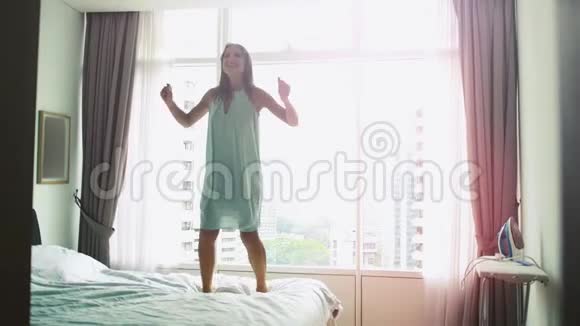 快乐的年轻女子跳在家里的床上跳舞在豪华公寓的床上玩有城市景观和镜头视频的预览图