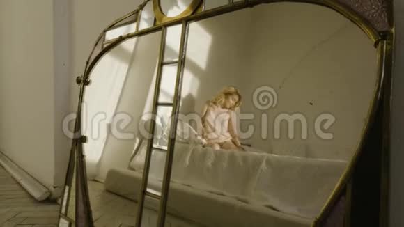 一个可爱的金发女孩在一张白色床上的睡衣弯曲她的腿行动镜子里的倒影视频的预览图