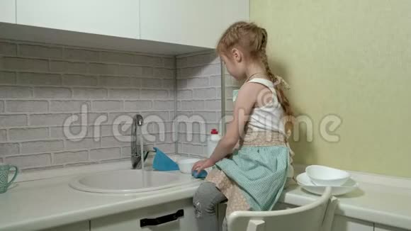穿着围裙的漂亮小女孩站在明亮的厨房里的椅子上在海绵上涂上洗涤剂有助于视频的预览图