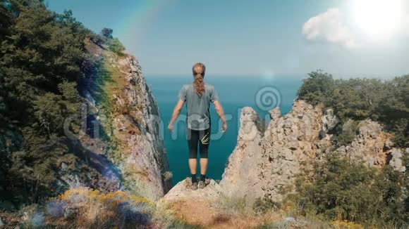 年轻的长发男子举起双手站在海面上一座山的顶端与美丽的山对着视频的预览图