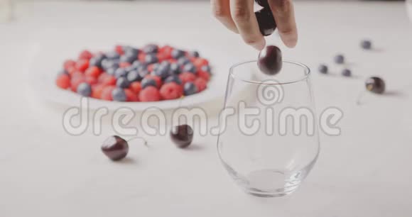 一个女人的手在杯子里装满成熟的红色浆果覆盆子樱桃蓝莓放在白色的桌子上模糊的视频的预览图