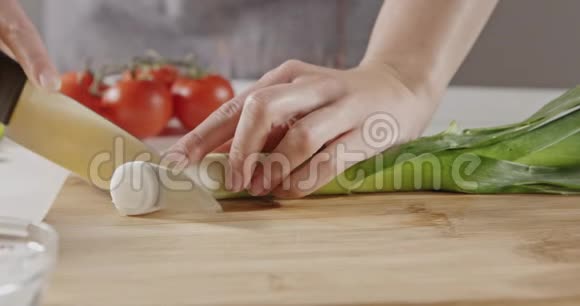 一个穿着围裙的女人把绿色的韭菜切在一个木切割板上在一张白色的厨房桌子上用红色做沙拉准备视频的预览图