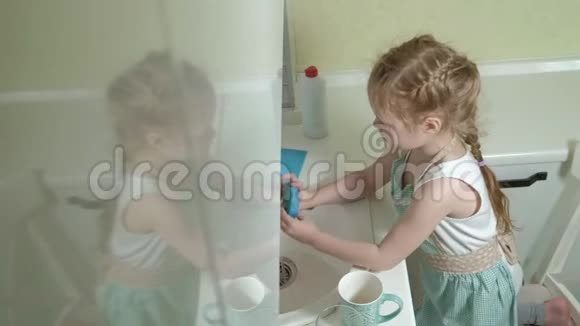 穿着围裙的漂亮小女孩站在椅子上在明亮的厨房洗碗帮助父母视频的预览图