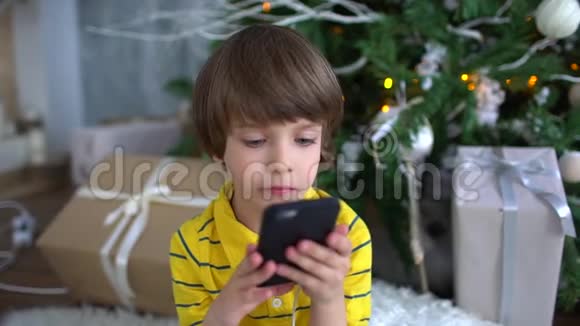 可爱的非常可爱的男孩坐在圣诞树的背景上玩智能手机慢动作儿童视频的预览图