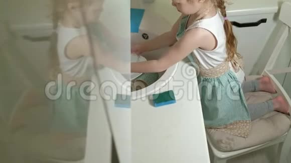 穿着围裙的漂亮小女孩站在椅子上在明亮的厨房洗碗帮助父母视频的预览图