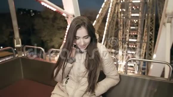 晚上年轻漂亮的女孩坐在摩天轮上在游乐园里用智能手机聊天视频的预览图