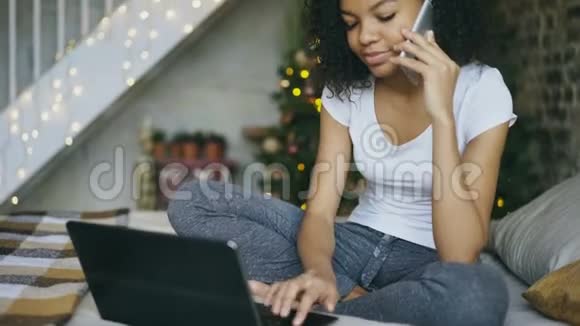 好奇的混血女孩在家里绕着圣诞树聊天智能手机和使用笔记本电脑视频的预览图