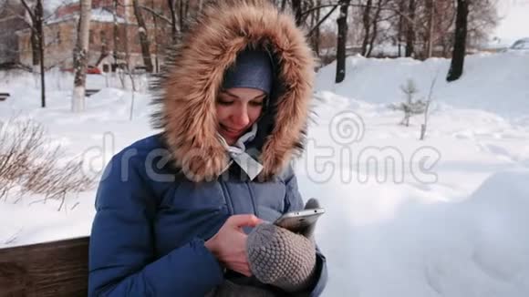 白天雪天特写女郎坐在长凳上在城市的冬季公园里浏览手机视频的预览图