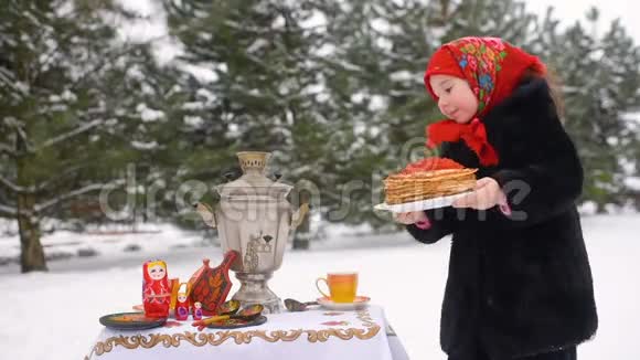 一个穿着毛皮大衣戴着俄罗斯风格围巾的小女孩在节日的桌子上放着一个盘子薄饼的盘子视频的预览图