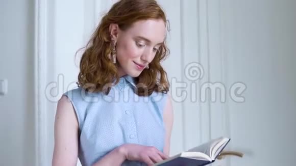 年轻的红头发女孩拿着一本书在白屋子里看书视频的预览图