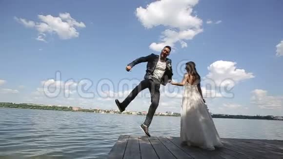 新郎站在新娘旁边跳跃可爱的新婚夫妇站在湖边慢动作视频的预览图