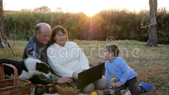 快乐的成熟夫妻与孙子和宠物一起野餐在背景日出时背景日出时带着笔记本电脑视频的预览图