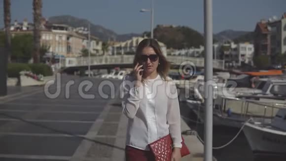 积极的商务女性戴着太阳镜在海的背景码头散步和打电话视频的预览图