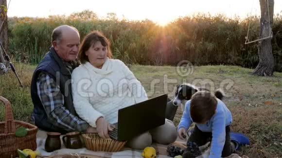 日出时野餐带着孙子和宠物的漂亮成熟夫妇坐在格子间手提电脑视频的预览图