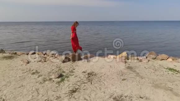 穿着红色长裙手舞足蹈的娇艳女子举起手在蓝色海水的壮观景色附近的大石头上视频的预览图