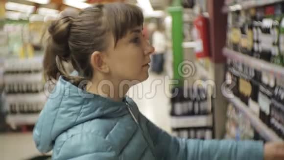超市里的女人年轻的白种人女士穿着蓝色夹克在瓶子上看着标签选择起泡葡萄酒视频的预览图