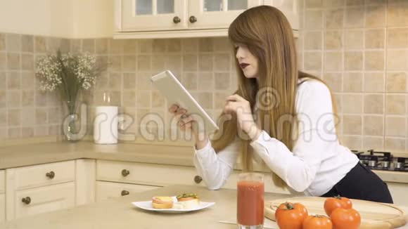 漂亮的女孩吃三明治在厨房里用平板电脑视频的预览图