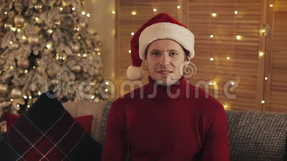 圣诞老人帽中的微笑魅力男人的看圣诞树背景上的相机慢动作视频的预览图